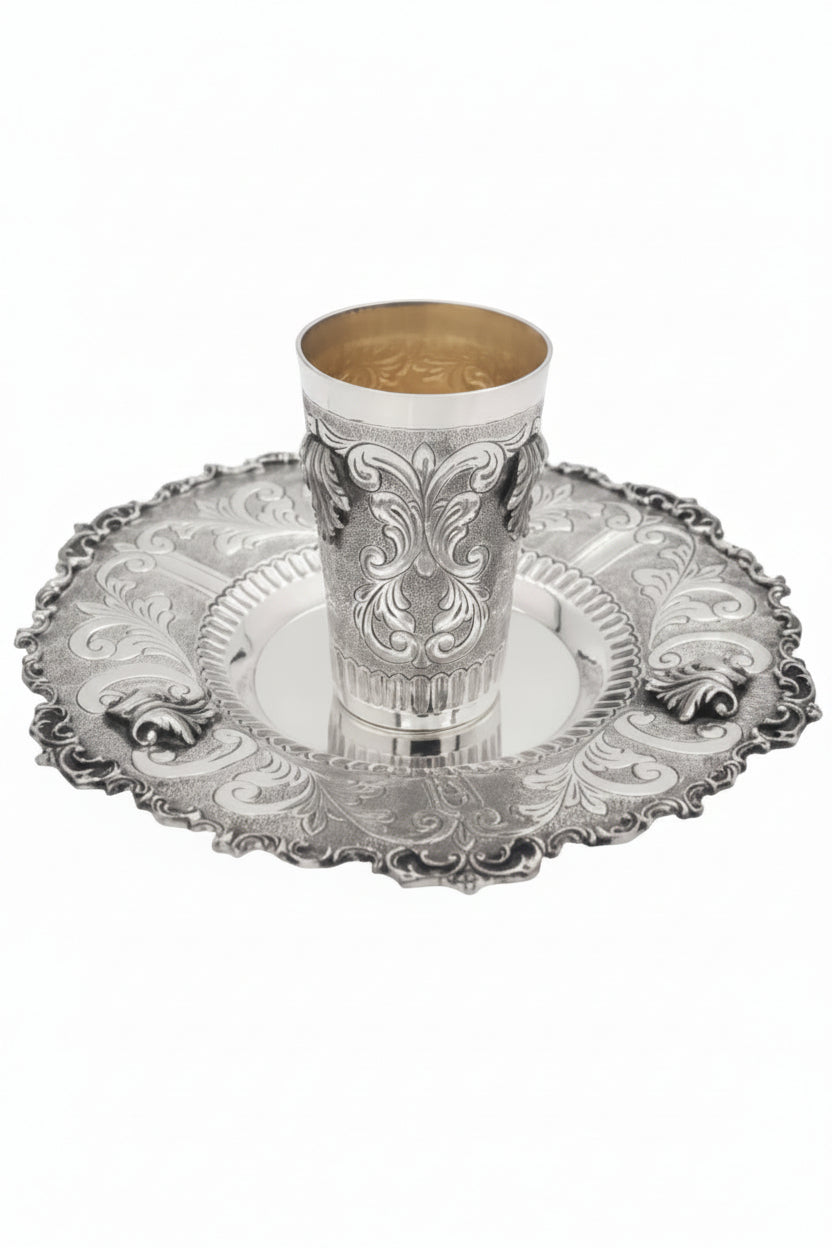 Silver kiddush cup with gold interior on an ornate silver tray against a white background